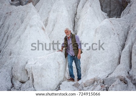 an old gray-haired tourist with a gray beard in jeans and a vest against a white cliff
