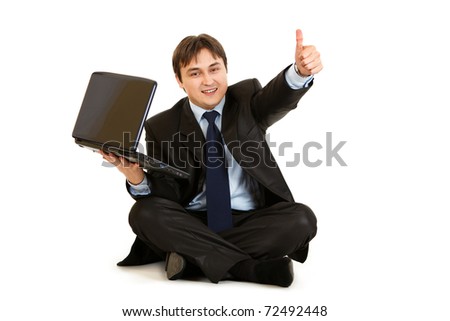 Smiling  businessman sitting on floor with laptop showing thumbs up gesture isolated on white