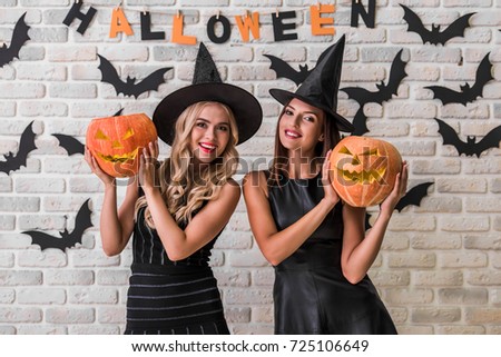 Beautiful girls in black dresses and witch hats are holding scary pumpkins, looking at camera and smiling on background decorated for Halloween