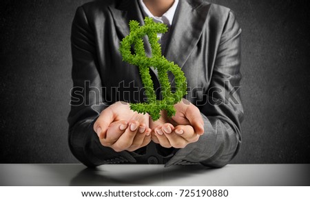 Cropped image of businessman in black suit presenting green plant in form of dollar symbol in hands with gray wall on background.