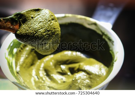 A spoonful of henna dye for hair, with a bowl of mixed henna paste in a blurred background.