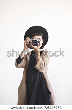 Vertical portrait of stylish young woman wears black dress, hat and beige linen coat. Girl takes pictures with her old film camera isolated on white wall background. 