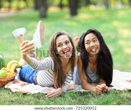 Caucasian and Asian young woman doing selfie and showing tongues. Funny girl friends having fun in free time in sunny park. International women friendship. Beautiful female models on picnic