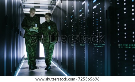 Two Military Men Walking in Data Center Corridor. One Uses Tablet Computer, They Have Discussion. Rows of Working Data Servers by their Sides. Royalty-Free Stock Photo #725365696