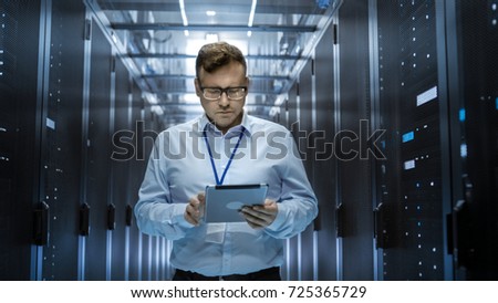 IT Technician Walks Through Rows of Server Racks in Data Center. Simultaneously He Works on a Tablet Computer. Royalty-Free Stock Photo #725365729