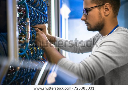 Side view portrait of young man wearing glasses connecting cables in server cabinet while working with supercomputer in data center Royalty-Free Stock Photo #725385031