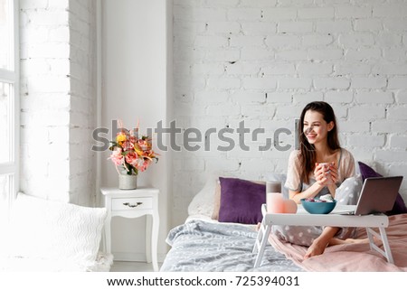 Joyful woman with cup of coffee in bed. Good morning with sweets and favorite drink combining with chatting on laptop or early working at home