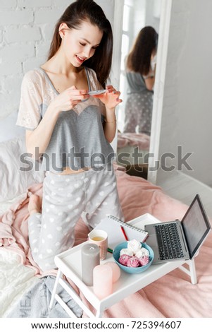 Smiling woman makes morning photo of working place by smartphone. Food photography during breakfast and chatting for social media. Zephyrs, cup of tea, laptop and writing book on small table