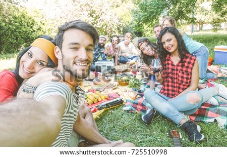 Group of friends taking a selfie in the park on a sunny day - Happy people having a picnic eating and drinking wine while taking photo with a mobile phone - Friendship, lifestyle, recreation concept 