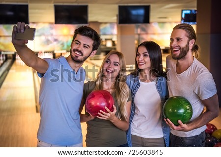Happy young friends are holding balls, doing selfie and smiling while playing bowling together