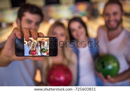 Happy young friends are holding balls, doing selfie and smiling while playing bowling together, phone in focus