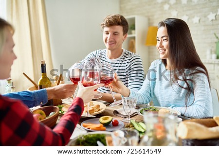 Group portrait of friends enjoying dinner together sitting at big table with delicious food , focus on young man and pretty Asian woman smiling happily clinking glasses celebrating holiday
