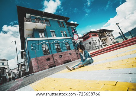 Wide-angle view of pensive black girl sitting on the paving stones right on the crosswalk of urban street on sunny autumn day with resort buildings in the background, Estosadok district, Sochi, Russia