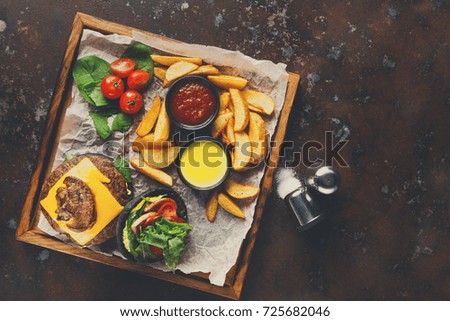 Take away burger menu on wooden tray at old rusty background, top view. Black bun cheeseburger with baked potato wedges and sauces set, fast food concept
