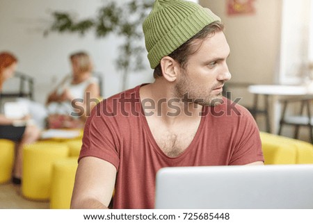 Indoor picture of attractive young bearded male wearing stylish hat sitting at coffee house in front of open laptop computer, working distantly, using free wifi with people talking in background