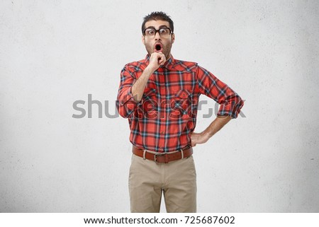 Picture of emotional adult Caucasian brunette guy with stubble wearing old fashioned spectacles and plaid shirt holding hand on chin, having shocked look as some bright mind crossed his mind