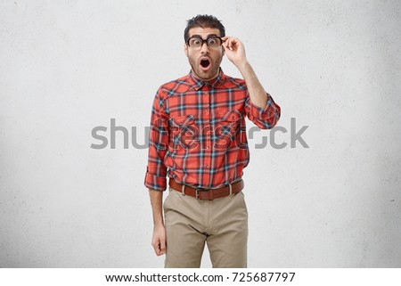 Horizontal shot of funny emotional male nerd in ridiculous clothes looking at camera in shock and terror with jaw dropped and eyes popped out as he sees some attractive amazing woman in front of him