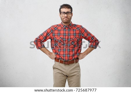 Portrait of furious bearded dark haired man geek looking ridiculous in old fashioned rectangular eyeglasses with thick lenses having serious or angry facial expression, keeping hands on his waist
