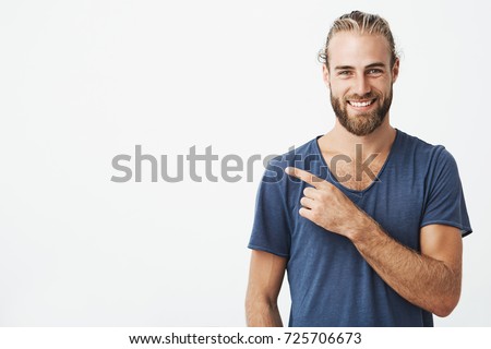 Happy beautiful bearded guy with good-looking hairstyle looking at camera, smiling and pointing aside with hand. Copy space. Royalty-Free Stock Photo #725706673
