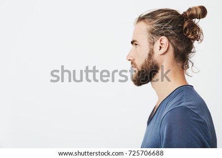 Close up of good-looking bearded hipster guy with hair in bun, in blue t-shirt standing in profile, looking aside, posing for photo. Royalty-Free Stock Photo #725706688