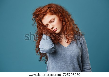 Portrait of unhappy frustrated girl with red wavy hair and freckles holding hand in hair with closed eyes having migraine or headache.