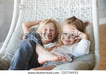 Picture of two siblings spending time together: blonde cute boy sitting in wicker hanging chair with his adorable little sister, both children looking at camera and smiling. Happy family and childhood