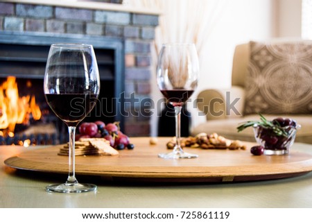 Cozy winter scene. Red wine and a snack platter with grapes, salted cracks, nuts and olives on a wooden board with a fire in a fireplace in the background. 