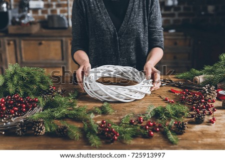 cropped view of florist making Christmas wreath of fir branches, decorative berries and pine cones at workplace