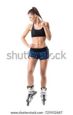 Confident fit sporty young woman in roller skates touching pony tail looking down. Full body length portrait isolated on white studio background.