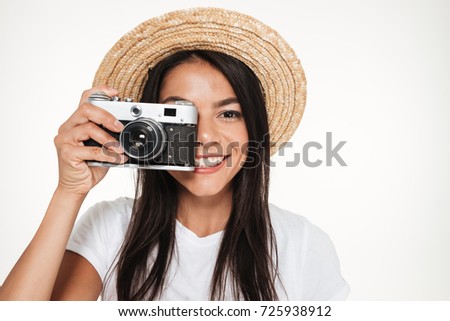 Close up portrait of a pretty young woman in hat standing and taking photo with a camera isolated over white background