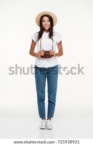 Full length portrait of a smiling young woman in hat standing and holding camera isolated over white background