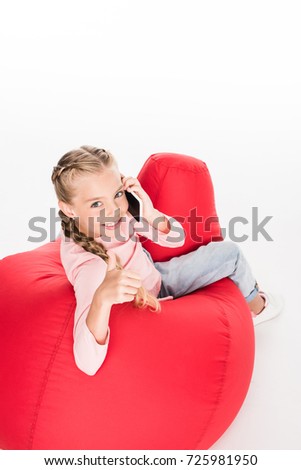 Young child sitting on a red bean bag with smartphone and showing thumb up, isolated on white