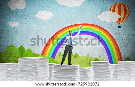 Businessman keeping hand with book up while standing on pile of paper documents with drawn landscape on background. Mixed media.