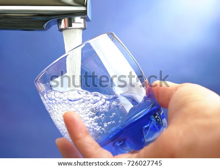 A hand holds a glass under a stream of clear transparent cold water from a tap on a blue background close-up of a macro.