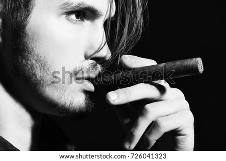 handsome bearded man with stylish hair on serious face smoking cigar in black studio background, close up