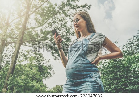 Young pregnant woman in denim overalls stands in park and uses smartphone. Girl using digital gadget, looking at smartphone's screen, chatting, blogging, checking email. Online marketing, education.