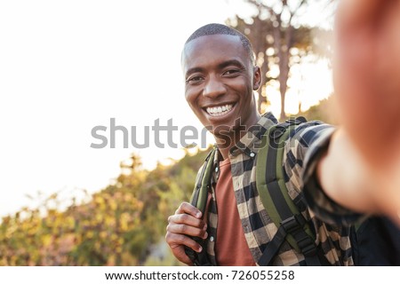 Smiling young African man in a backpack taking selfies while hiking alone up a trail on a sunny day