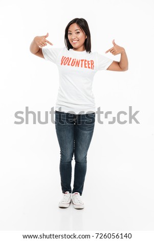Full length portrait of a happy young asian woman in volunteer t-shirt standing and pointing two fingers while looking at camera isolated over white background