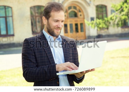 Business expert. Confident handsome man with beard in shirt hold laptop and smiling while standing against university background. 