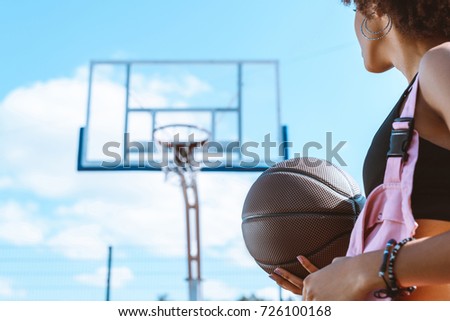 african-american woman in sports bra and pink overalls holding a basketball ball at sports court