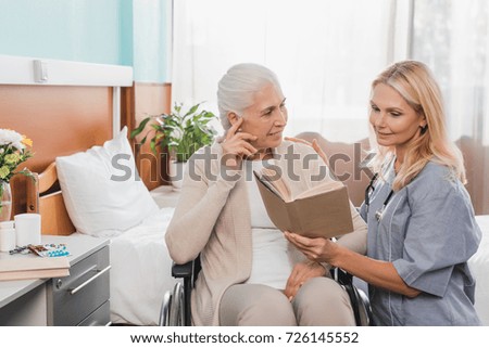 smiling nurse reading book to senior patient with hearing aid sitting in wheelchair 