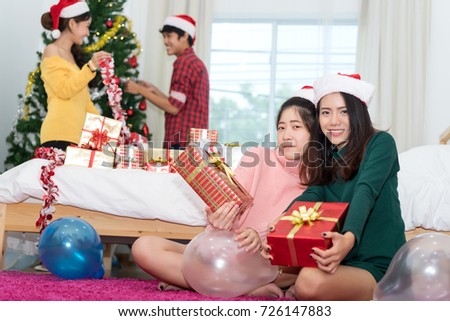 Young Asian group man and women together teenager friend with santa hat holding gift box surprise christmas holidays and  christmas tree background in room at home relaxing celebrating new year