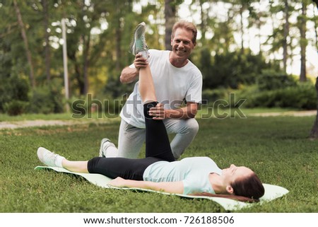 A man helps his girl to do stretching. She is lying on a rug for yoga, a man is holding her leg. Behind them is the summer park