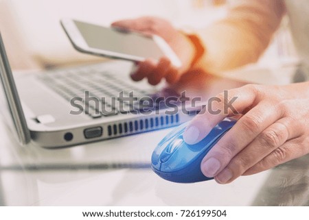 Hand on computer mouse and smartphone in the other. notebook in background in desk