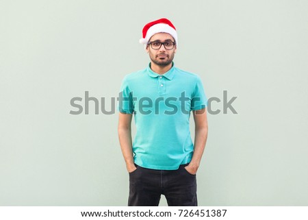 Happiness bearded man in Christmas red hat and black glasses standing near gray wall and looking at camera. Studio shot