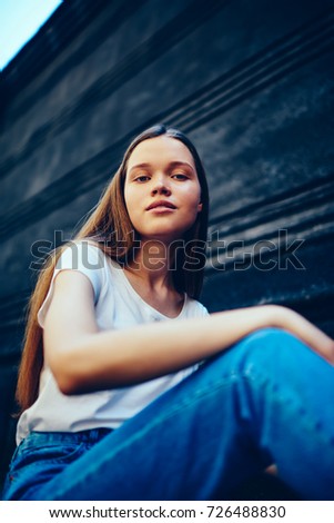 Portrait of casually dressed attractive hipster girl  looking at camera sitting outdoors near wall background,charming female with natural beauty makeup in fashionable apparel posing for photo