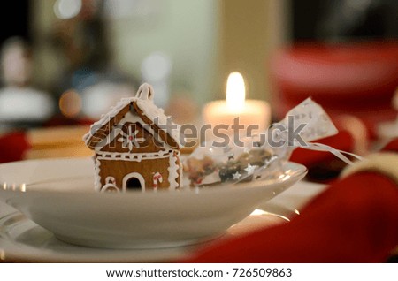 A picture of a festive table setting with red Christmas theme. A small handmade ginger house sits inside a soup plate as a decoration and a white candle is lit in the background. 