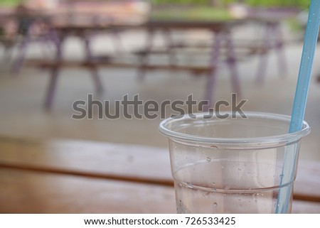The empty plastic glass of water with condensation inside the a blue straw with background with a dining table, which is placed inside the cafeteria.