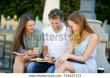 Three students are preparing for exams. Two girls and a guy sitting on a bench and talking animatedly. In the hands of their notes and their tablet.