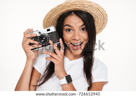Close up portrait of a cheerful young woman in hat standing and holding a camera isolated over white background